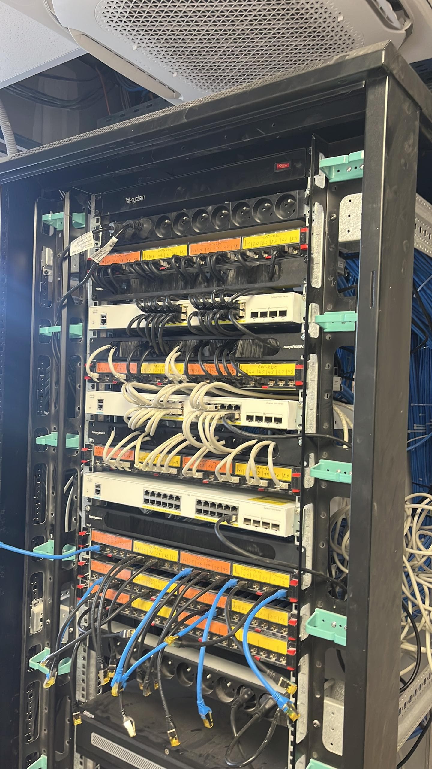 Baie de brassage pendant le travail : panneaux étiquetés à la main, switches Cisco Catalyst 1200, câbles à réorganiser en cours de classement.