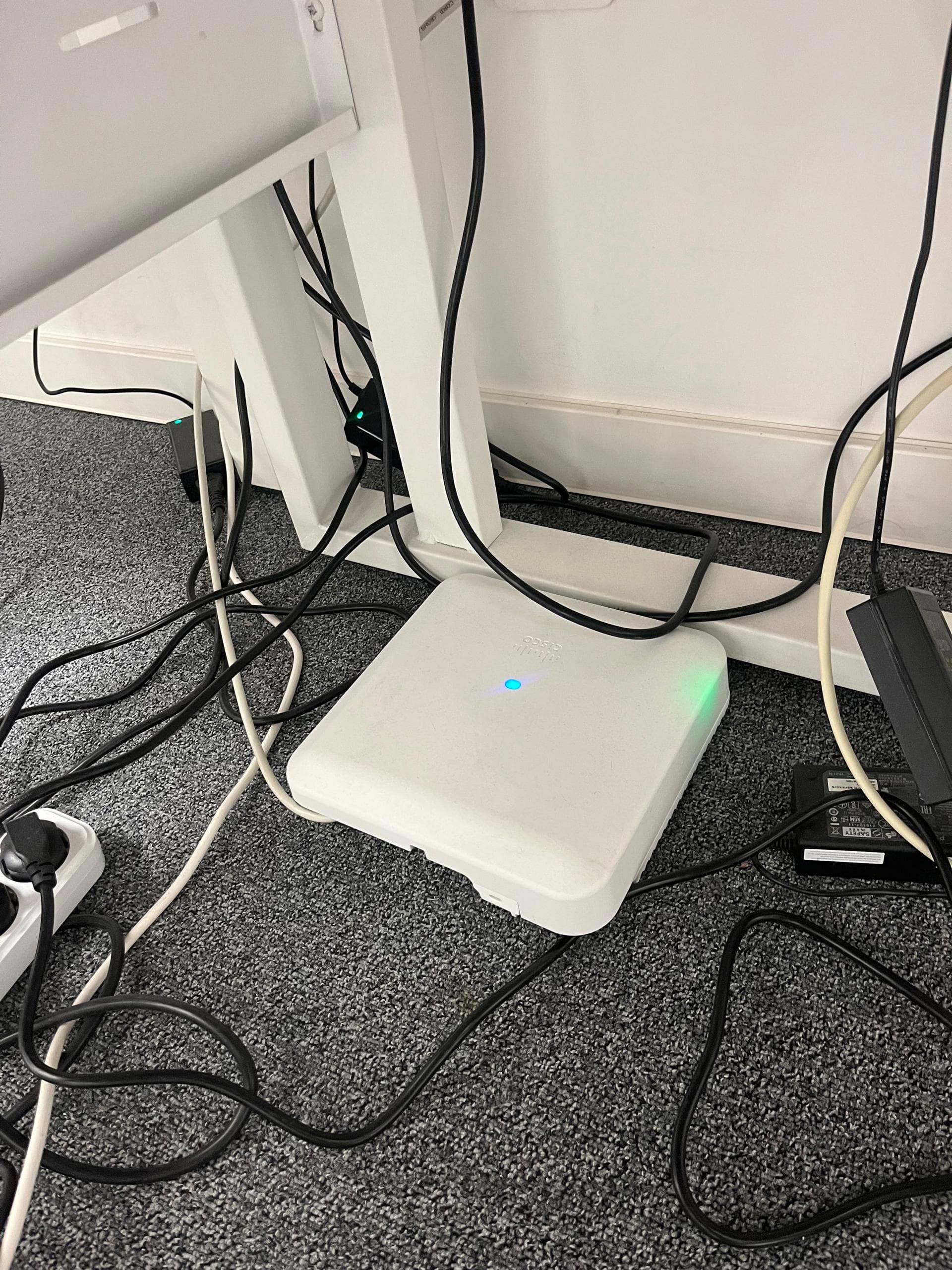 Point d'accès Cisco posé au pied du bureau, relié par ses câbles réseau et alimentation, voyants allumés.